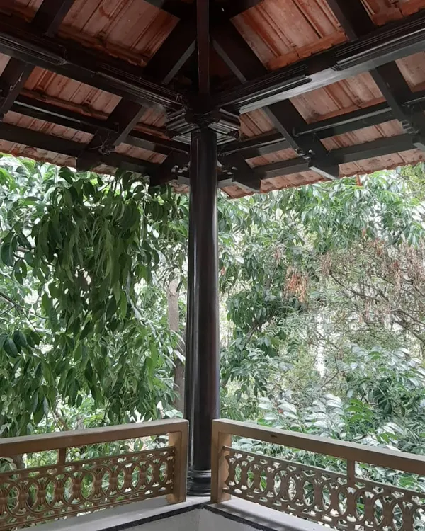 A view from a restored balcony, featuring traditional wooden pillars and intricately carved railings set against a lush green backdrop. The terracotta tile roof completes the authentic, vernacular feel.