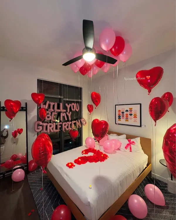I can help you ask the big question with a beautiful surprise setup. This room features 'WILL YOU BE MY GIRLFRIEND' foil balloons, a mix of red and pink latex and heart-shaped foil balloons, and a bed covered in rose petals to create the perfect romantic proposal setting.