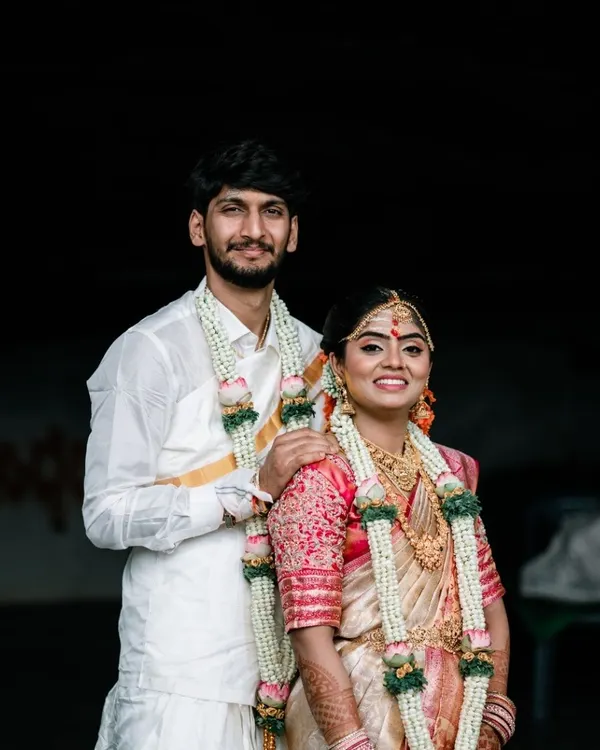 A classic portrait of a South Indian couple on their wedding day. Their traditional white and gold attire, along with the floral garlands, looks beautiful against the simple, dark background.