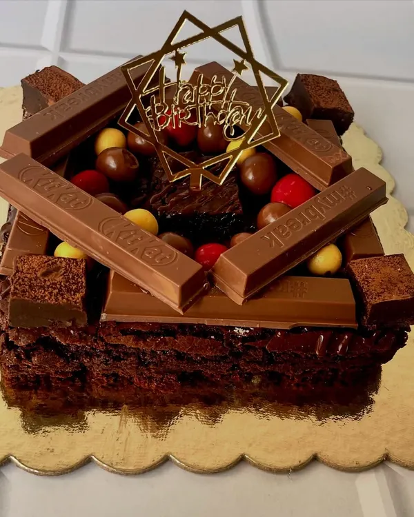 Decadence, redefined. This overloaded brownie cake is topped with KitKat bars, colorful gems, and brownie chunks, finished with a "Happy Birthday" topper. It's an irresistible treat for any chocolate lover's celebration.