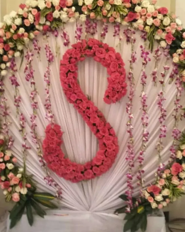 Personalized decorations make any event feel special. This backdrop features a large circular frame of white and pink roses, with the initial 'S' made entirely of bright pink roses as the centerpiece. It's a great idea for birthdays, anniversaries, or engagement parties.