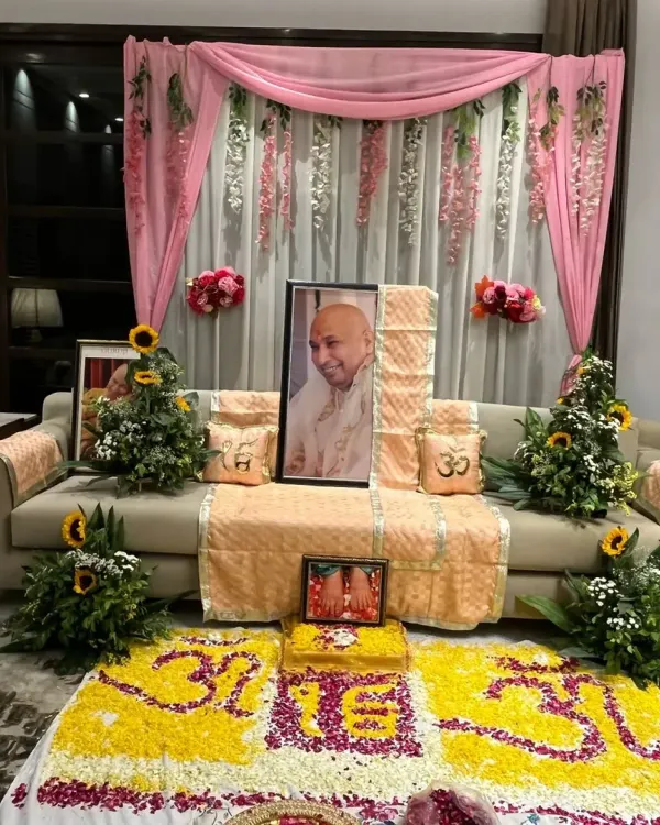 An elegant and serene setup for a Guruji satsang. The decor features a beautiful floral rangoli, sunflower arrangements, and a pink drape backdrop with hanging wisteria to create a divine ambiance.