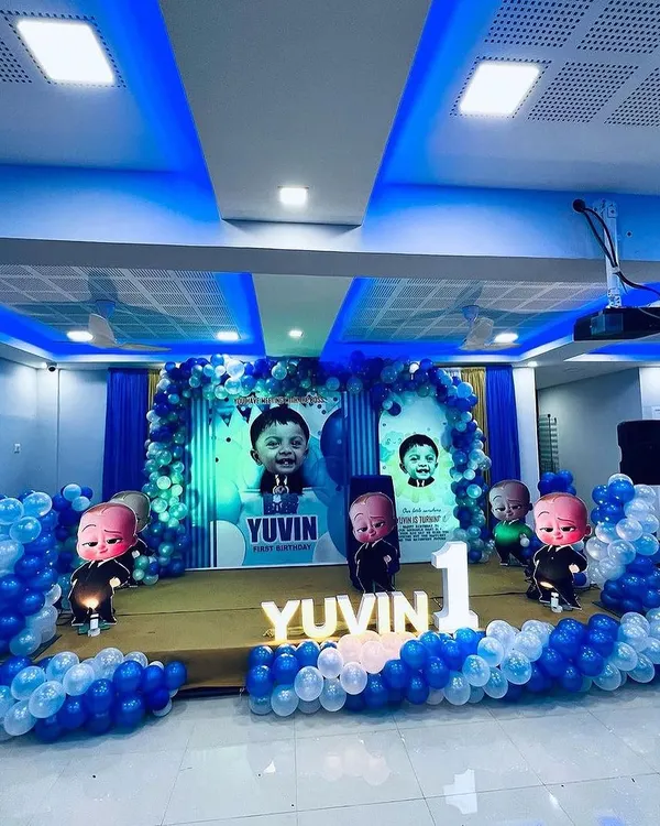 A complete Boss Baby themed stage setup for a first birthday. This includes personalized backdrops with the child's photo, multiple Boss Baby character cutouts, and a blue and white balloon arch.