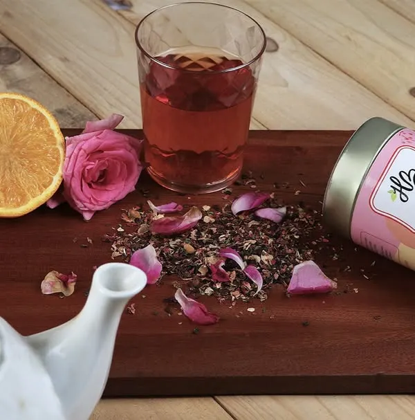A lifestyle composition for a rose and lemon herbal tea brand. The image features the product tin, a brewed cup, and fresh ingredients like rose petals and an orange slice on a rustic wooden board.