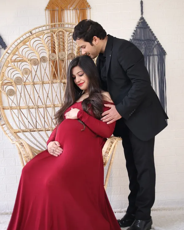 A romantic studio portrait of a couple with a peacock chair prop. The partner gently kisses the mother-to-be's head as she cradles her bump.