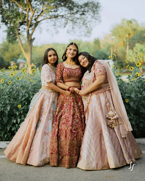 A bride stands happily with her two sisters, all in beautiful lehengas, in a sun-drenched garden.