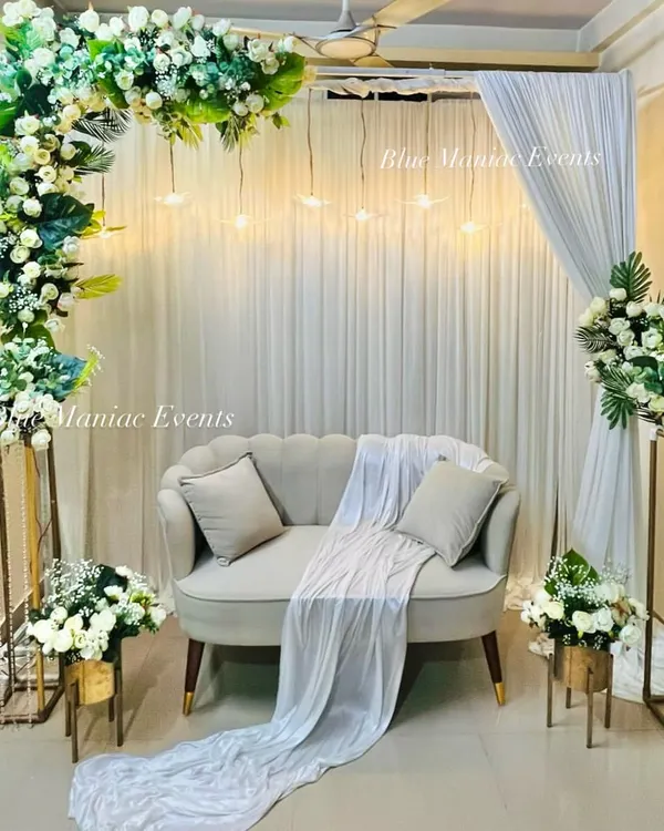 A breathtaking Roka setup featuring a lush floral arch of white roses and green leaves, with hanging lights creating a magical glow. The decor is complemented by floral stands and a classic sofa.