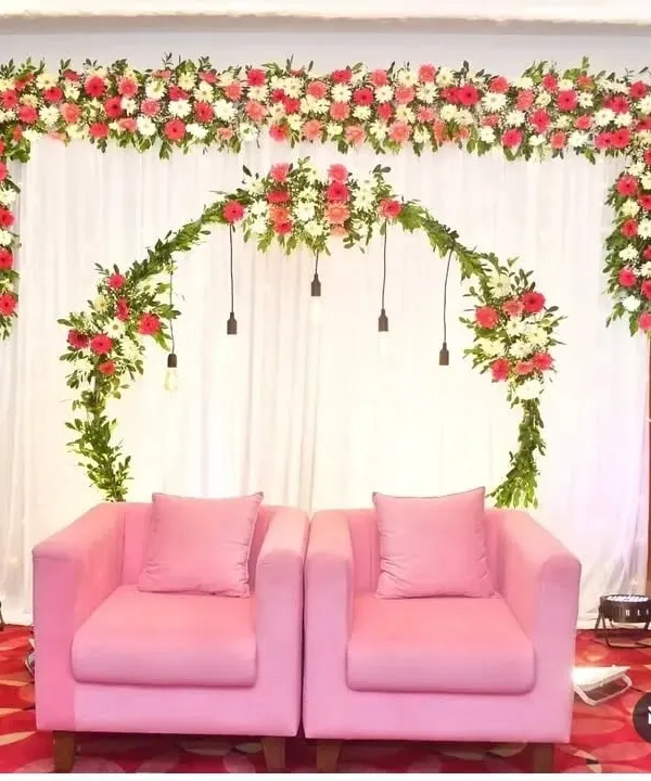 A beautiful stage backdrop for an engagement ceremony featuring a clean white curtain. The decor is highlighted by a lush floral arch of red and white flowers with hanging Edison bulbs, perfectly framing the two pink armchairs for the couple.