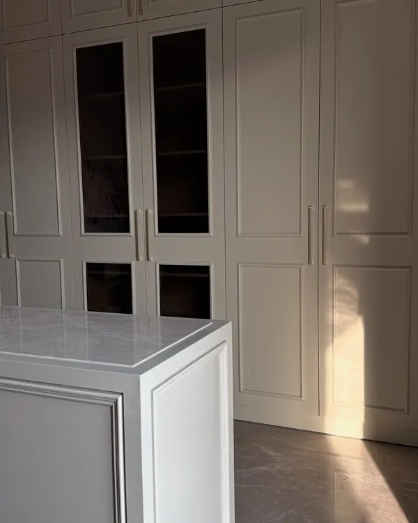A stunning walk-in closet featuring floor-to-ceiling off-white wardrobes with classic paneling and a matching central island with a marble top. The design exudes elegance and offers ample storage.