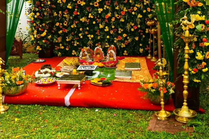 A pooja laid out on a red carpet in a garden in front of a wall of flowers: four small pots dressed in garlands, plates of fruit, flowers and betel, a steel box, and tall brass lamps at the sides