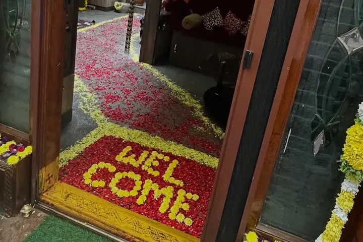 A welcome rangoli at a home's front door: WELCOME spelt in yellow flowers on a bed of red rose petals edged in yellow, with a path of scattered red petals leading inside and white and yellow flower garlands on the door frame