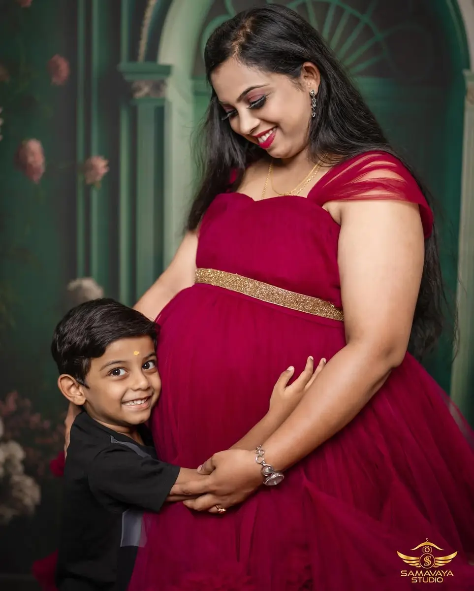 A smiling young boy hugs his pregnant mother's bump, her in a deep red gown, in a studio