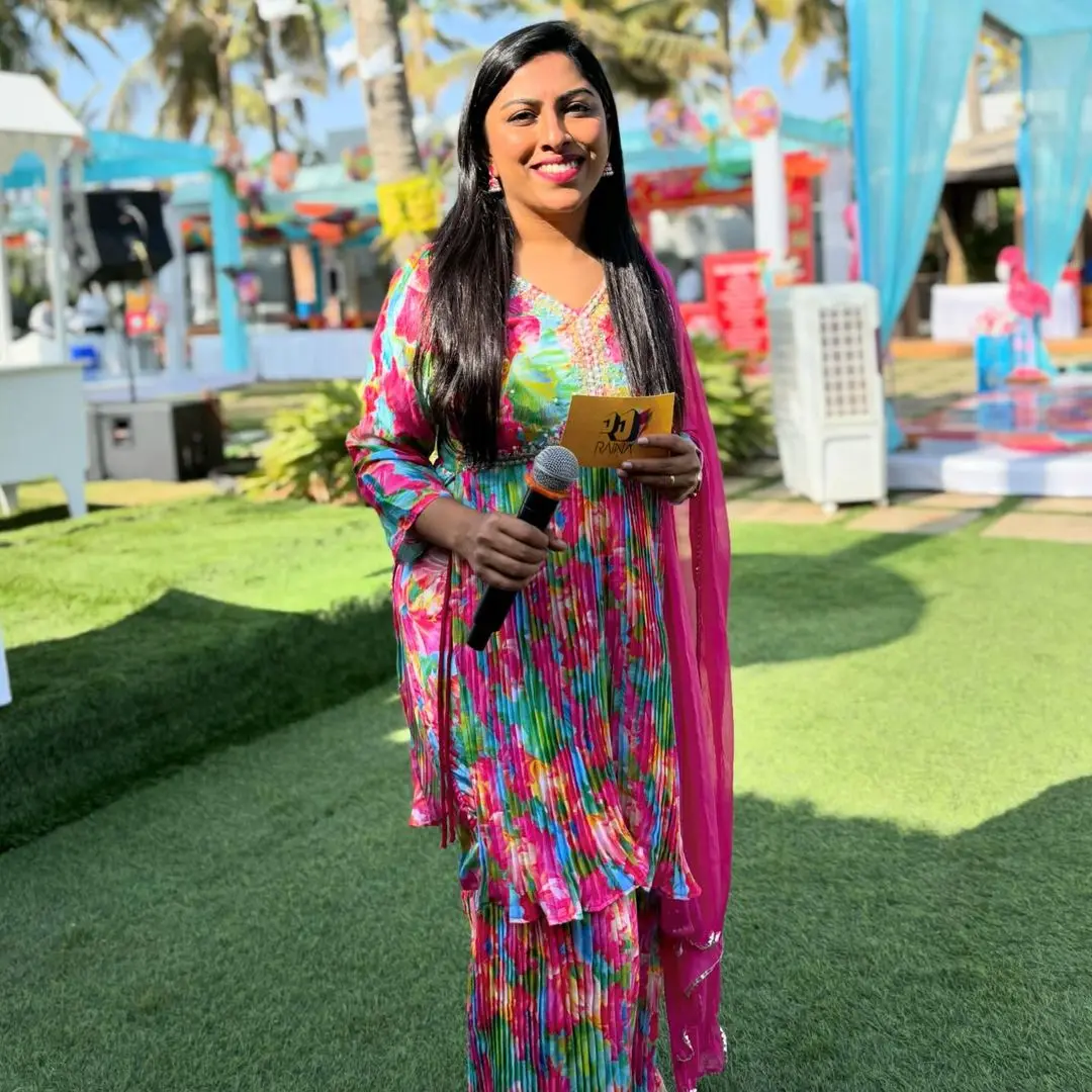 Raina Dsouza at an outdoor mehendi on a lawn, in a bright floral kurta and pink dupatta, a microphone in one hand and a gold cue card in the other, with a black speaker on a stand and a sound case behind her on the left