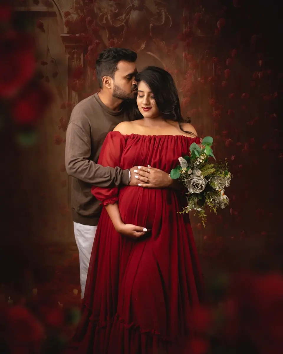 A pregnant woman in a red off-shoulder gown holds a bouquet of grey roses while her husband in a brown sweater kisses her head, against a dark backdrop of climbing red roses