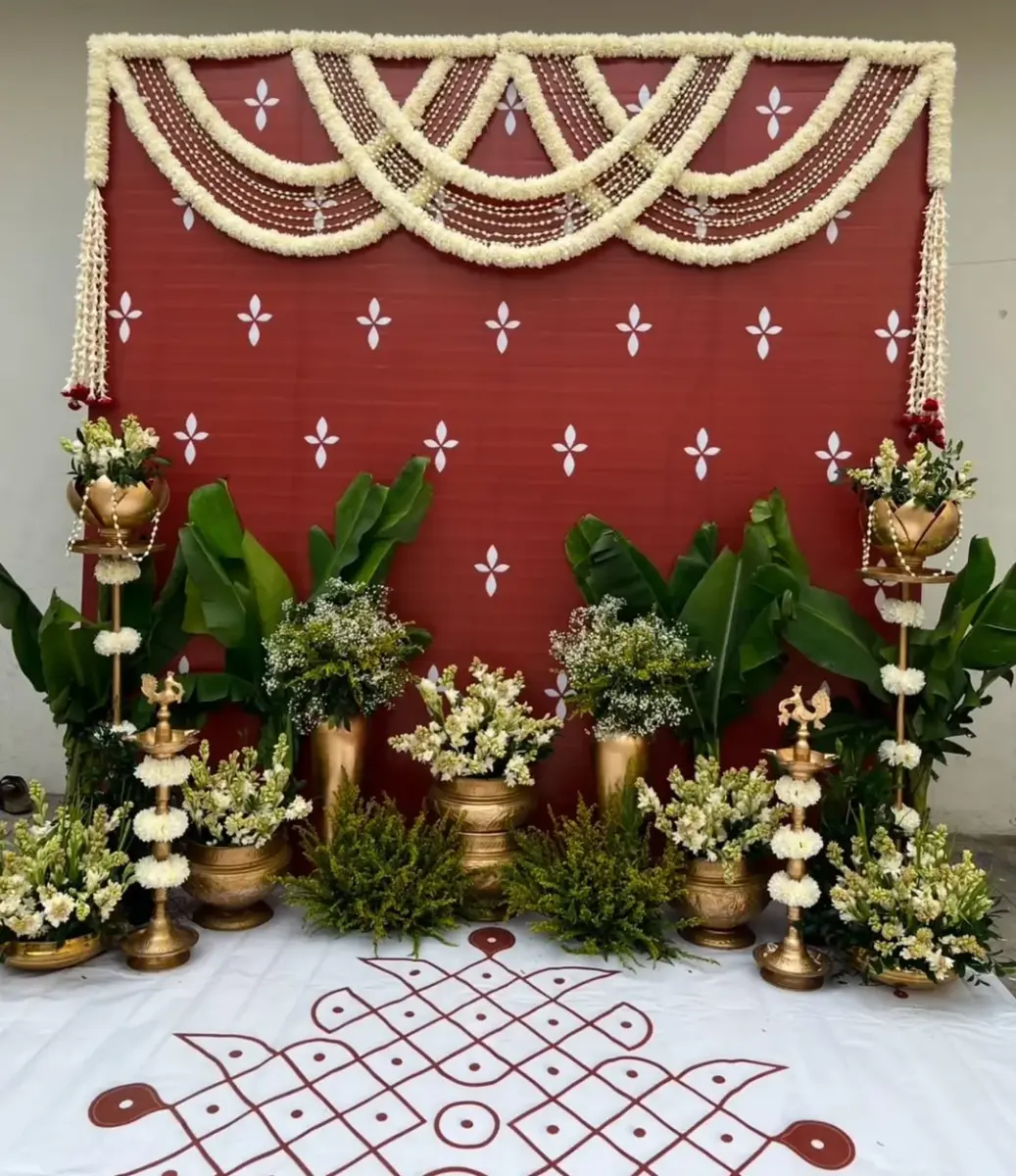 A red kolam of loops and dots on a white cloth spread over the floor, in front of a red backdrop with white motifs, swags of white flowers, banana plants, brass pots of white flowers and brass lamp stands