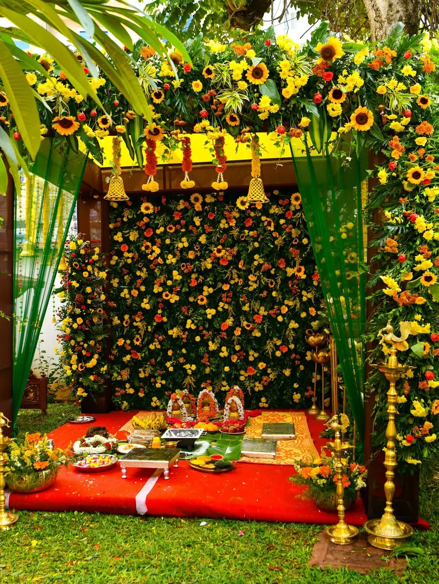 A mandap in a home garden with a roof of sunflowers, marigold and gerbera, a wall of the same flowers behind, green curtains, tall brass lamps, brass pots of flowers, four kalasha with garlands and offerings on a red carpet
