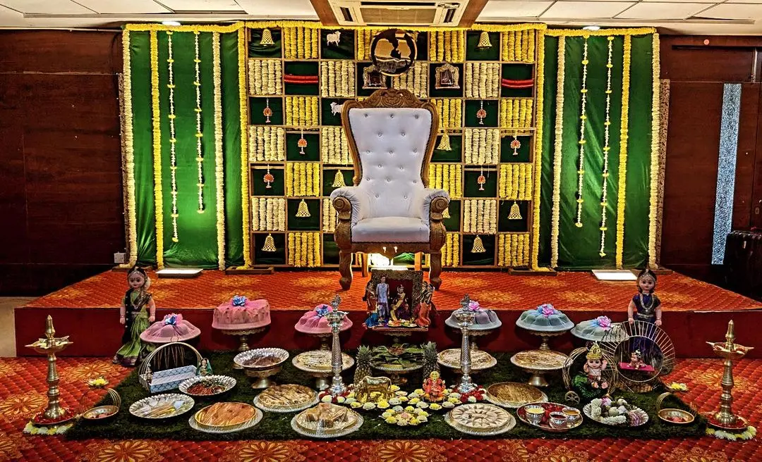 A seemantham stage in a hall: a green and marigold backdrop with a checker of yellow flowers, a white high-backed seat in the middle, and plates of fruit, sweets and dolls laid out on a green mat in front, with tall brass lamps at each end